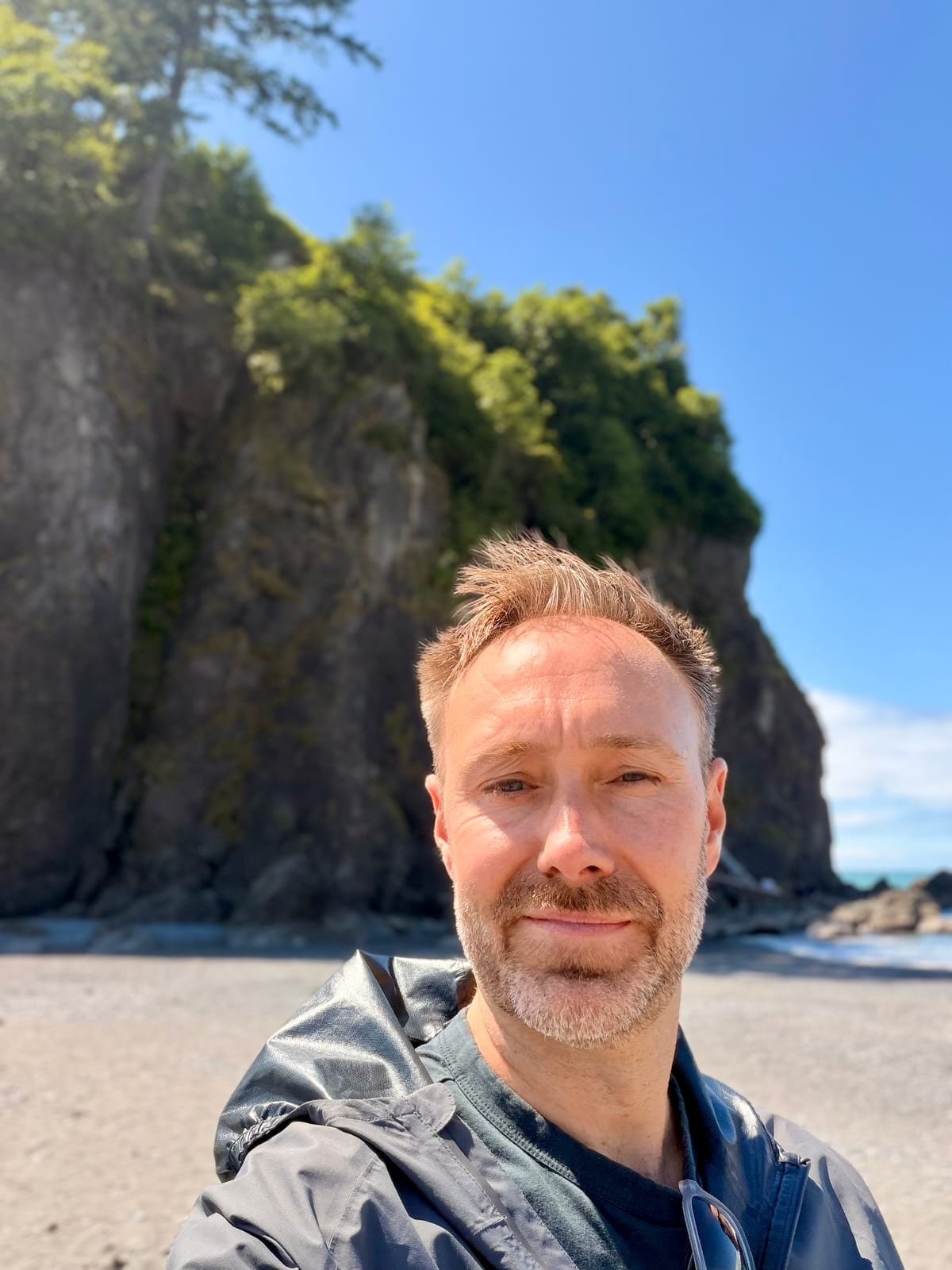 Ken taking a selfie on a sunny beach in front of a forested sea cliff