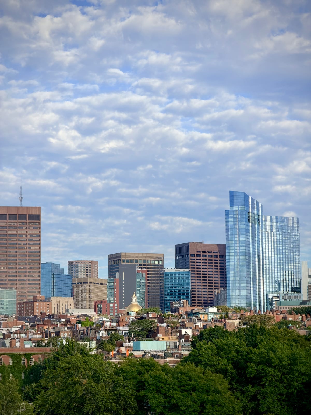 A gold dome among Boston's brick rooftops, green trees, and modern towers beneath a sky of rippled clouds