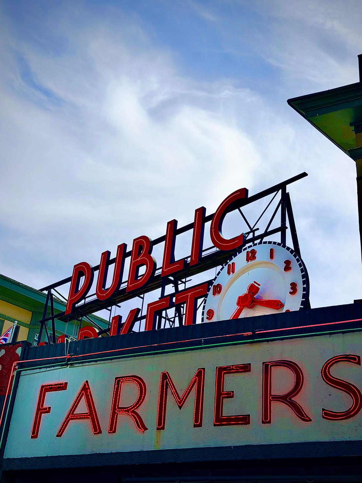 Red Public Market lettering and a clock above a glowing Farmers neon sign beneath a cloudy Seattle sky