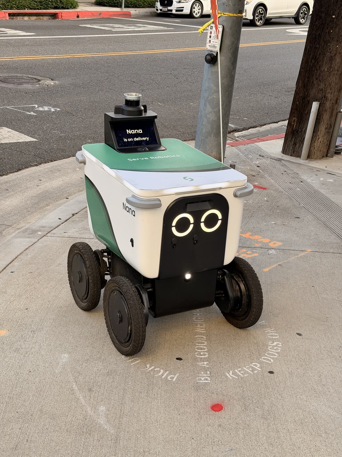 A small green and white Serve Robotics delivery robot named Nana waiting at a West Hollywood crosswalk