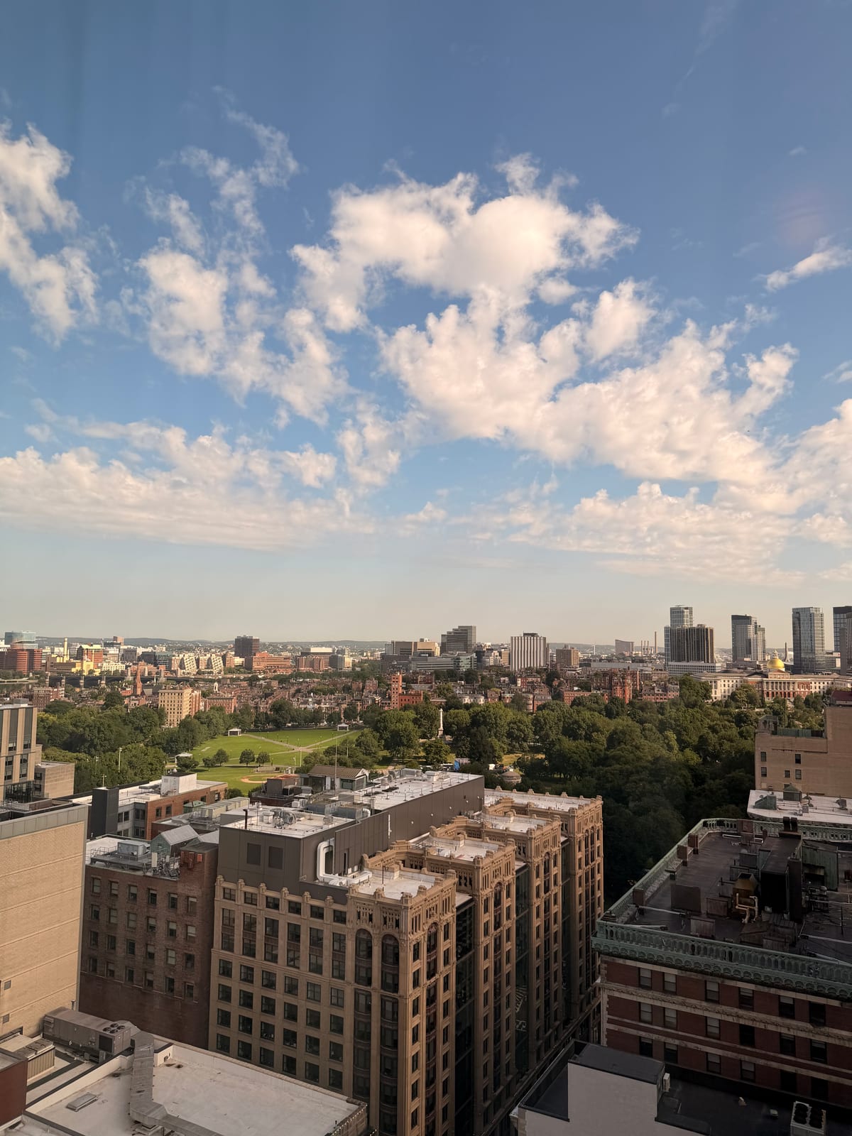A wide view over downtown Boston, the Common, and the Massachusetts State House beneath soft summer clouds