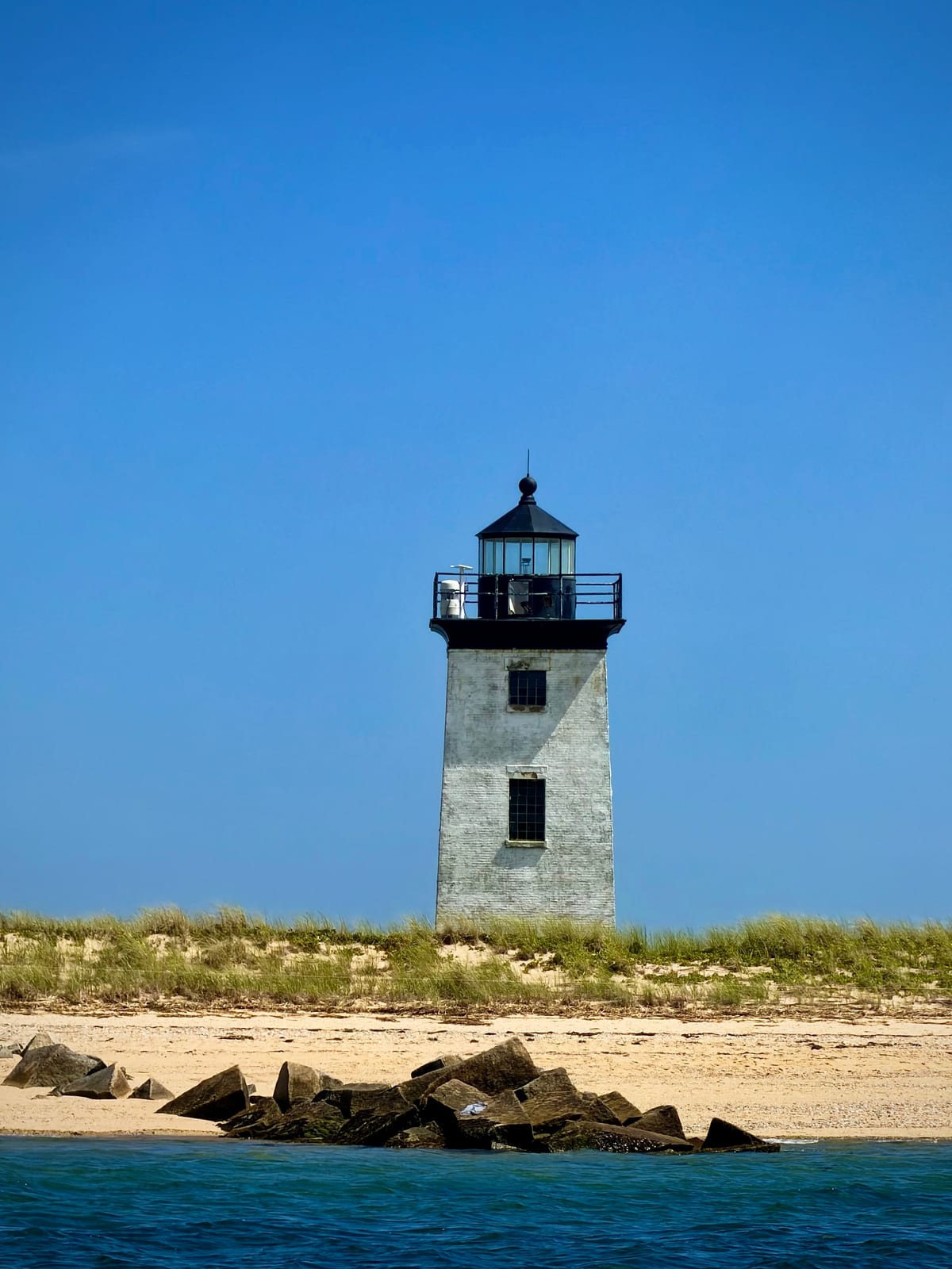 A white lighthouse rising above dunes beside bright blue water