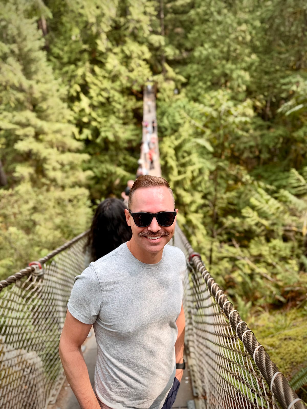 A smiling man standing on a suspension bridge above a forested canyon
