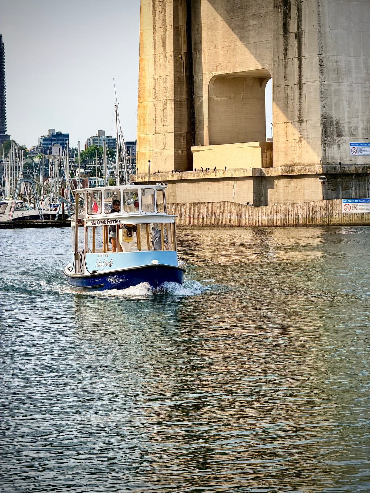 A tiny blue-and-white False Creek ferry approaching beneath a large concrete bridge