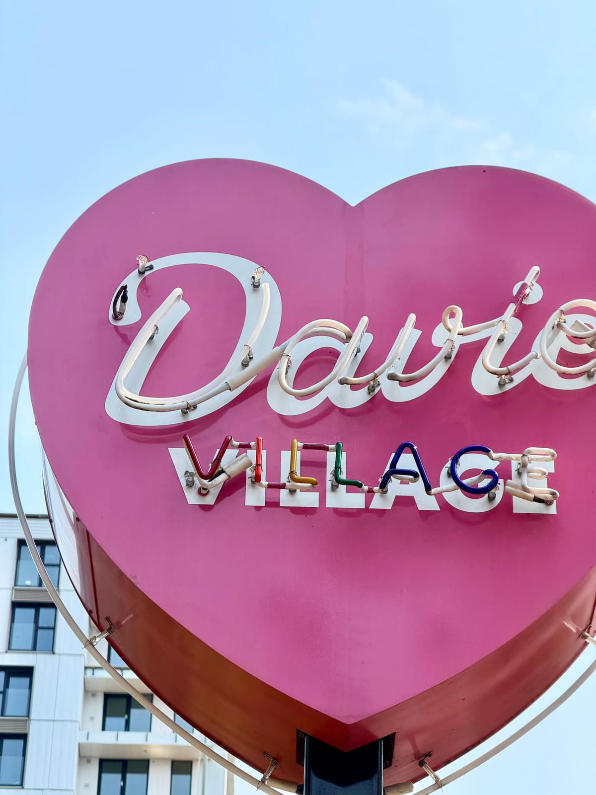 A pink heart-shaped Davie Village neon sign against a blue sky
