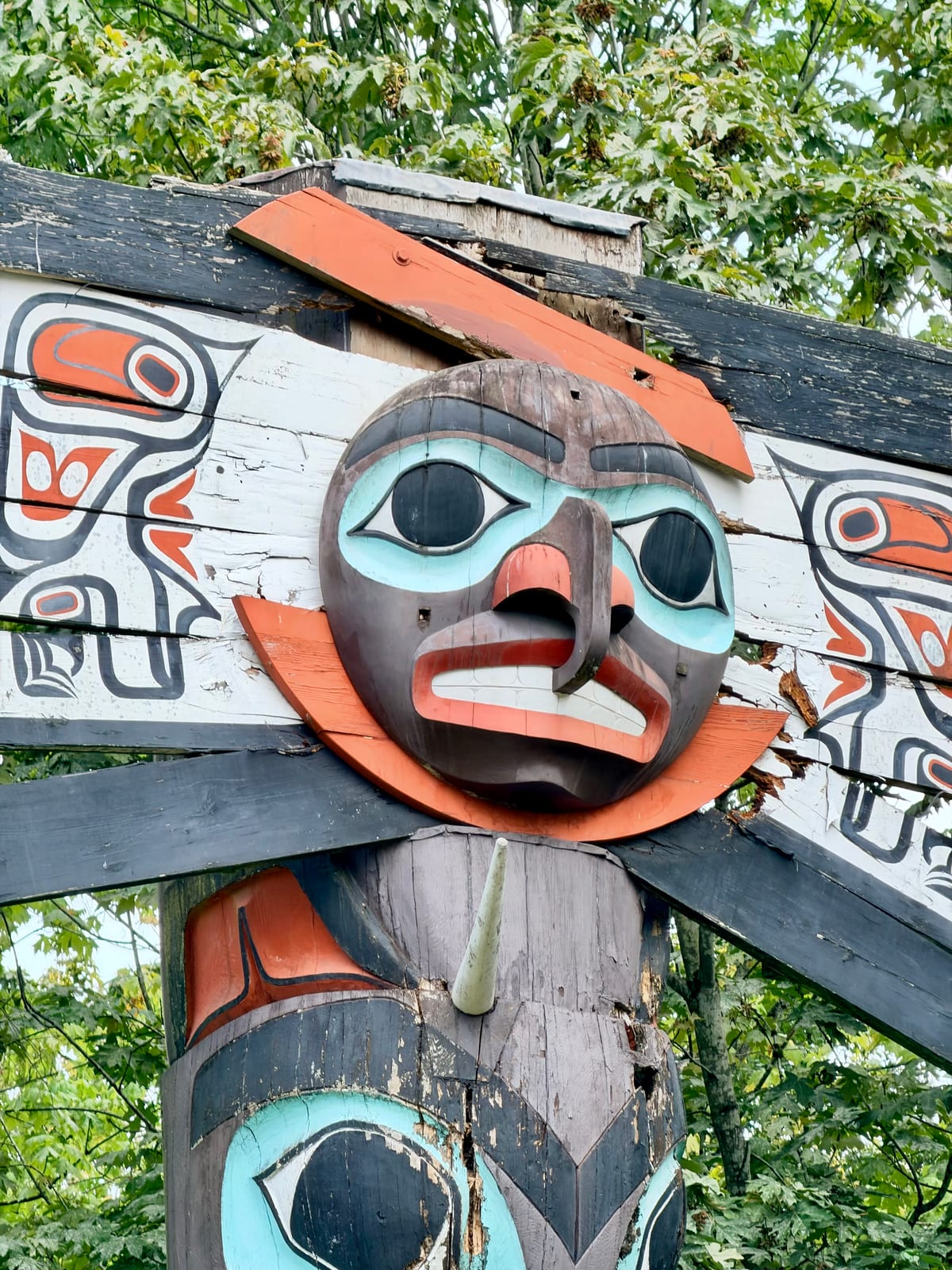 A close-up of a weathered carved wooden face painted turquoise, orange, black, and white