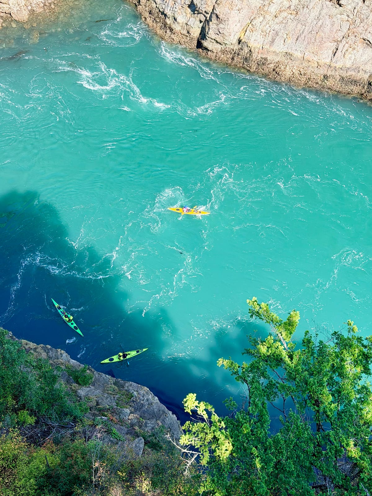 Three colorful kayaks navigating swirling turquoise water below Deception Pass Bridge